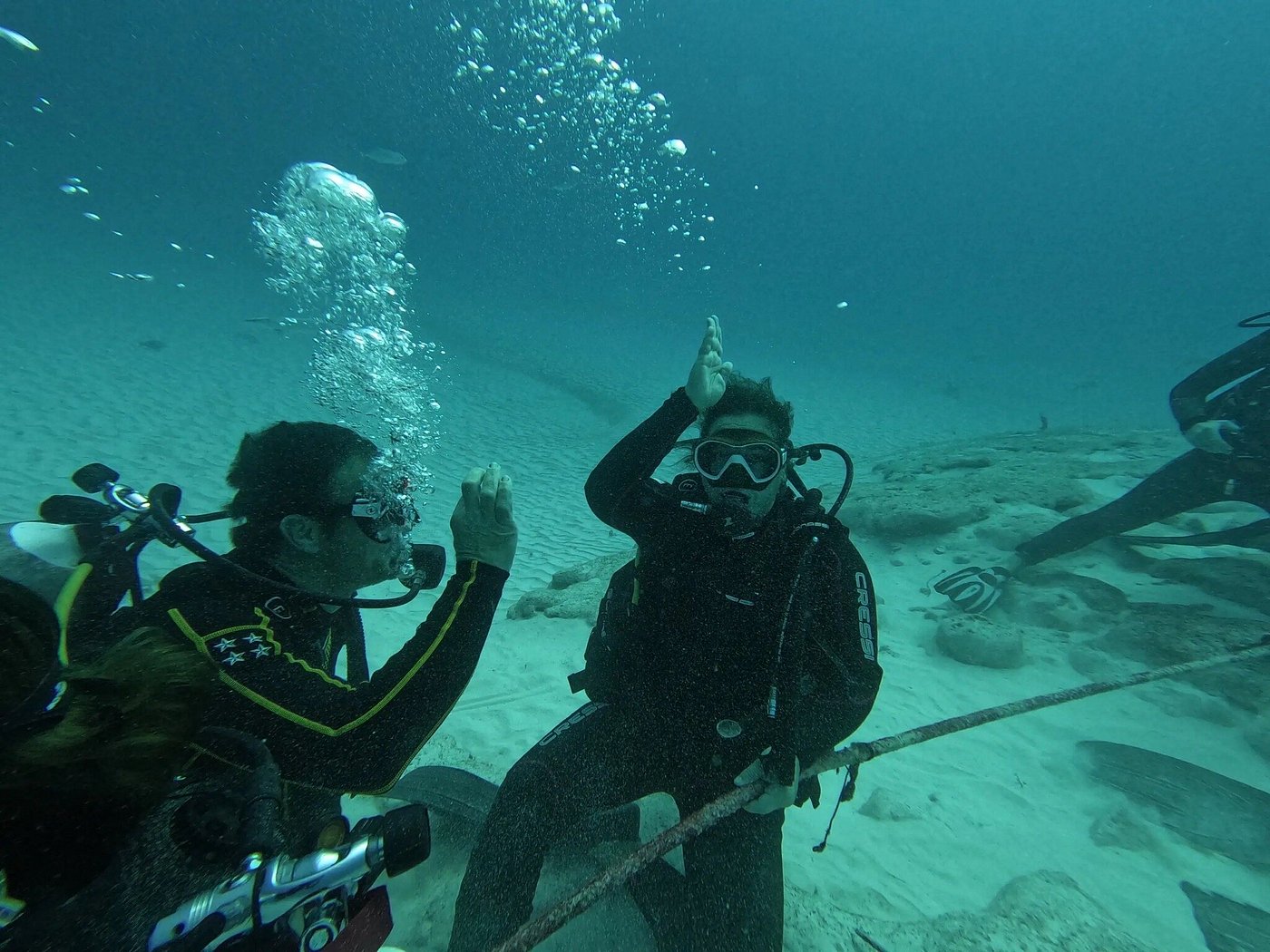 Two scuba divers underwater giving shark signals over a sandy ocean floor.